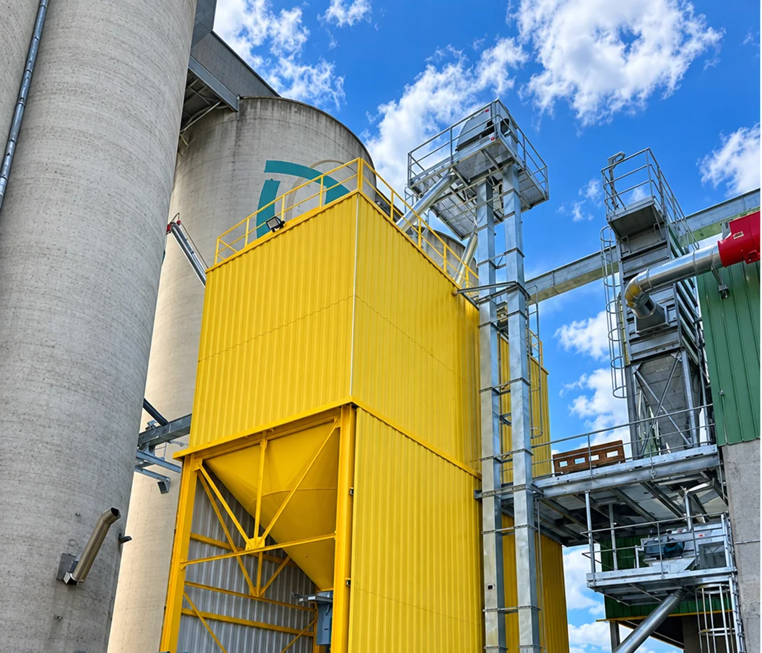 Grande cellule peinte en jaune chrome, trémie et gaine métallique, silos béton en arrière-plan.