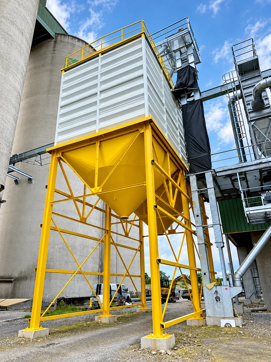 Trémie et charpente peintes en jaune chrome devant une cellule blanche et des silos béton.